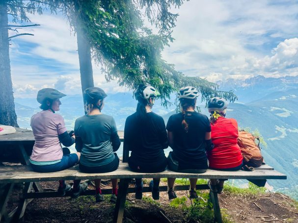 E-MTB Camp Dolomiti 2026 Teilnehmerinnen sitzen bei einer Tourpause auf einer Holzbank an der Titschenwarte und blicken auf das Panorama der Dolomiten.