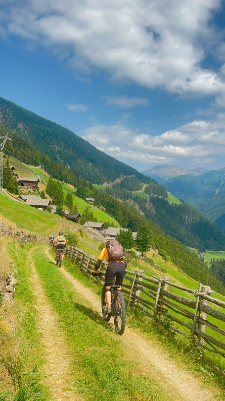 E-MTB Camp Ultental 2026 Zwei E-MTB-Fahrerinnen fahren auf einem schmalen Wiesenweg durch die Berglandschaft des Ultentals in Südtirol.