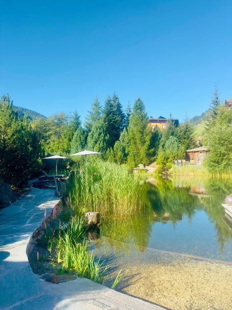 E-MTB Camp Ultental 2026 Naturteich des AROSEA Life Balance Hotels im Ultental mit Schilf, Bäumen und einem Sitzplatz.