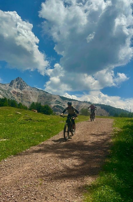 E-MTB Camp Dolomiti 2026 Zwei E-MTB-Fahrerinnen fahren auf einem breiten Schotterweg in Richtung der Felsberge beim Camp Dolomiti.