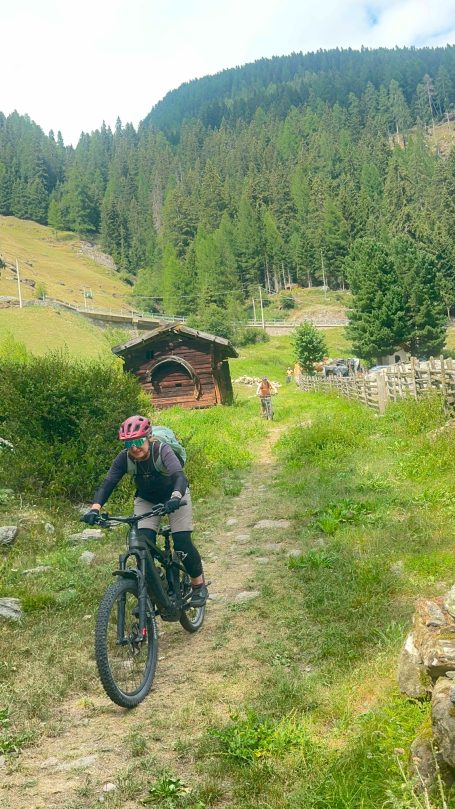 E-MTB Camp Ultental 2026 E-MTB-Fahrerin fährt auf einem schmalen Bergtrail an einer Almhütte und Weidezäunen im Ultental in Südtirol vorbei.