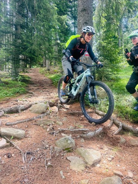 E-MTB Camp Dolomiti 2026 E-MTB-Fahrerin bewältigt eine felsige Waldpassage, während eine Begleiterin am Rand Hilfestellung gibt.