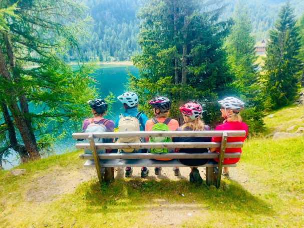E-MTB Camp Ultental 2026 Fünf E-MTB-Fahrerinnen sitzen auf einer Bank und blicken beim Camp im Ultental auf einen Bergsee und bewaldete Hänge.