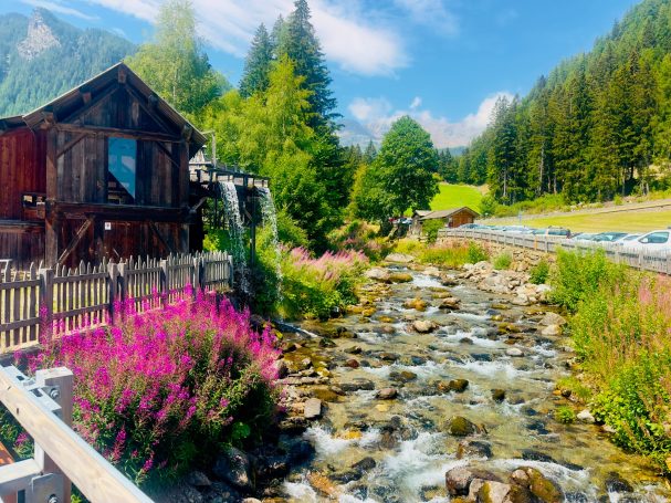 E-MTB Camp Ultental 2026 Bergbach mit Holzhaus, Blumen und bewaldeten Hängen entlang der Route des E-MTB-Camps im Ultental.