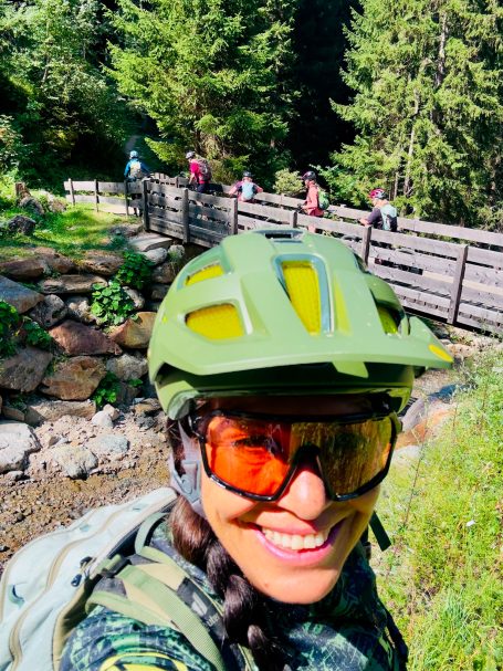 E-MTB Camp Ultental 2026 E-MTB-Fahrerin macht ein Selfie vor einer Holzbrücke, während die Gruppe mit Bikes einen Bach im Ultental in Südtirol überquert.