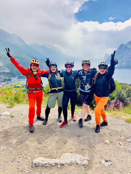 E-MTB Camp Gardasee 2026 Fünf Teilnehmerinnen stehen mit erhobenen Armen am Monte Brione mit dem Gardasee und den Bergen im Hintergrund.
